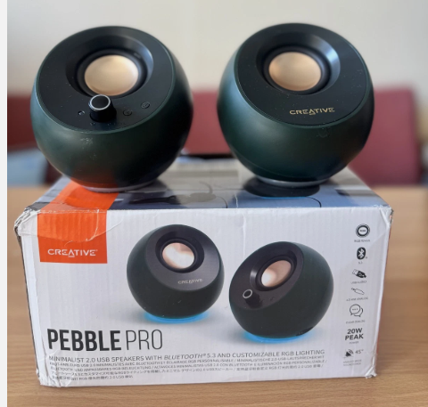 Two dark green Creative Pebble Pro wireless speakers displayed on top of their original white and orange retail box, set on a wooden surface.