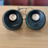 A pair of Creative dome-shaped computer speakers in a dark green color, placed on a light wood tabletop with their connecting cable visible behind them.