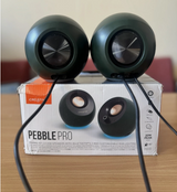A pair of dark green Pebble Pro Bluetooth speakers with black cables, resting on top of their white product packaging box on a wooden table.